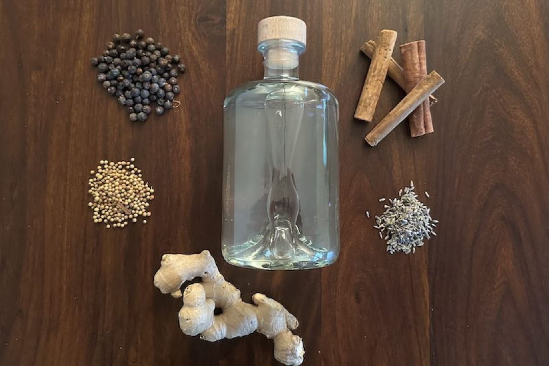 Klare Gin-Flasche auf dunkler Holzoberfläche, umgeben von klassischen Botanicals wie Wacholderbeeren, Koriandersamen, Zimt, Lavendel und Ingwer.