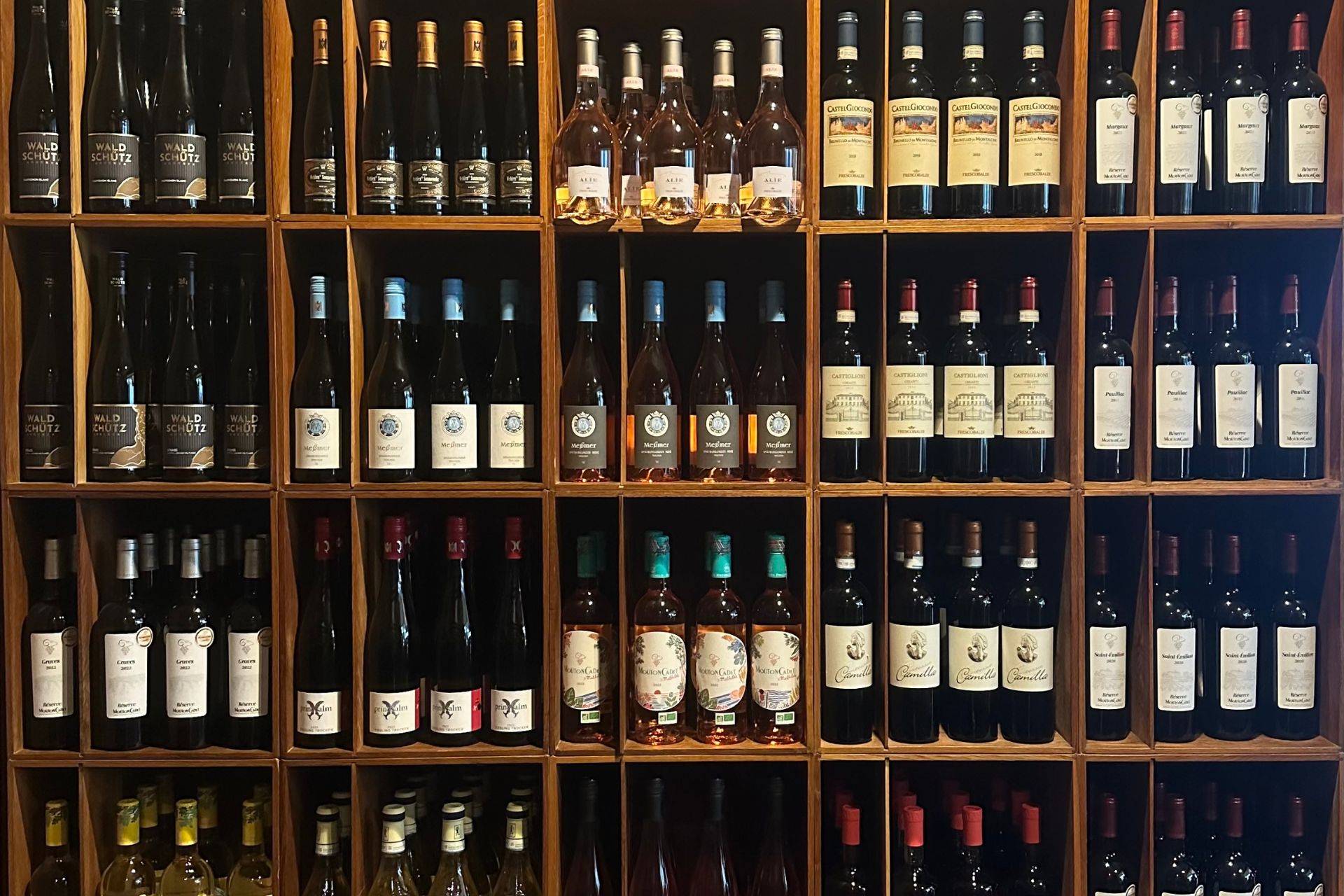Ein großes Weinregal mit verschiedenen Flaschen Rot-, Weiß- und Roséwein, sorgfältig sortiert in Holzfächern. Die Vielfalt der Weine spiegelt die Auswahl für eine Weinprobe wider, bei der verschiedene Rebsorten und Anbaugebiete entdeckt werden können.